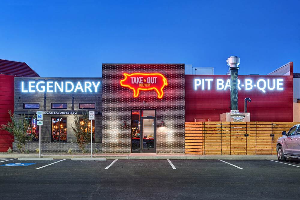 Exterior of Famous Dave’s BBQ with bold signage reading “Legendary Pit Bar-B-Que” and a neon pig highlighting “Take Out Delivery”—a modern, eye-catching facade promoting America’s favorite BBQ experience with dine-in and takeout options.