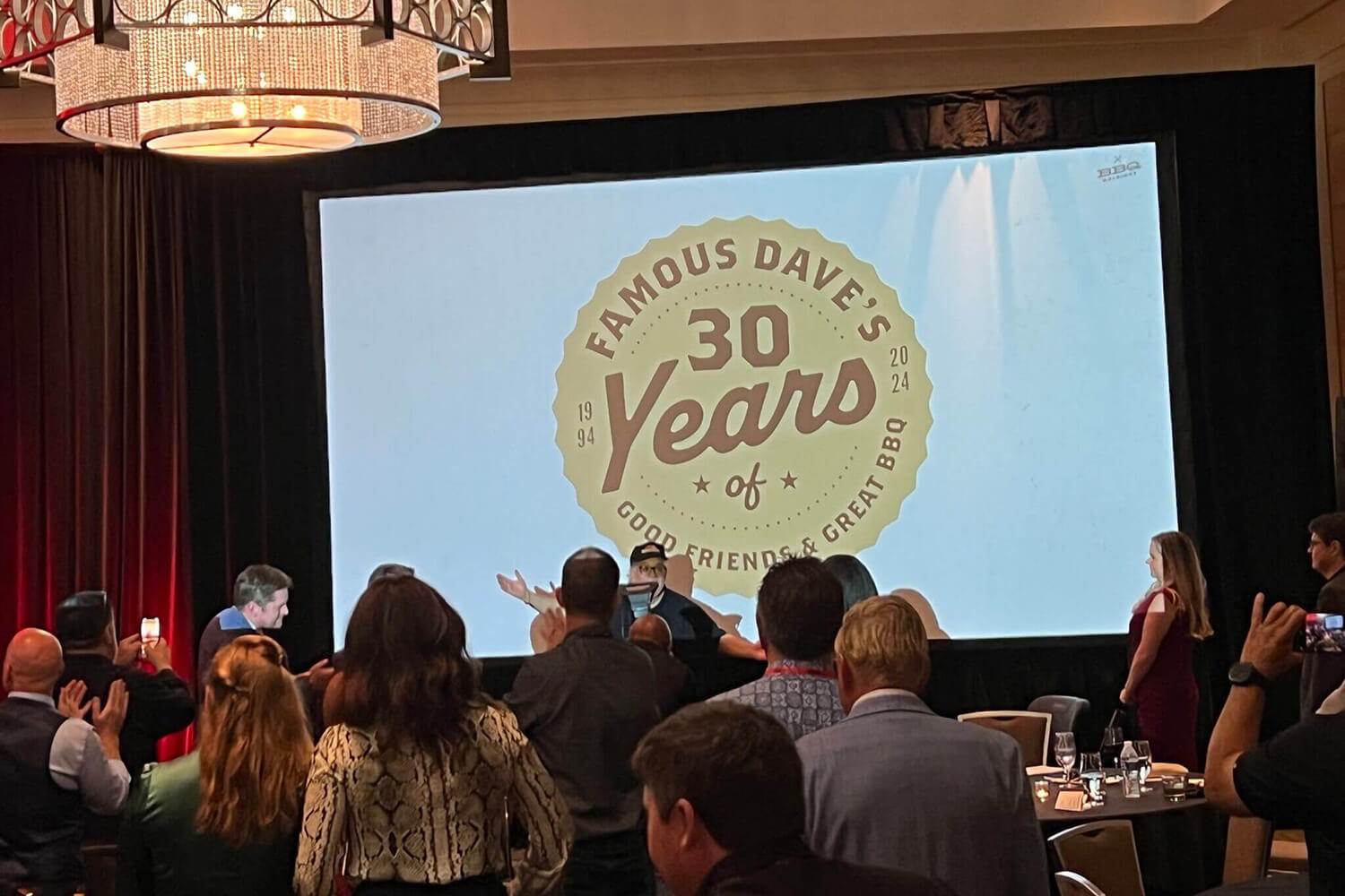 Attendees, including members of Triple A Group, gather and applaud during the 2024 BBQ Holdings Conference in Dallas, TX. A large screen displays “Famous Dave’s 30 Years of Good Friends & Great BBQ,” celebrating the brand’s 30th anniversary.