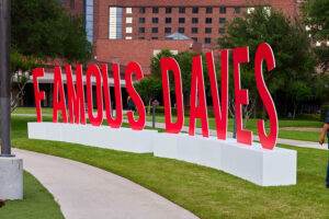 A bold red “Famous Dave’s” sign displayed on a white base outdoors at the 2024 BBQ Holdings Conference in Dallas, TX, attended by the Triple A Group. The backdrop features a brick building and a landscaped walkway.