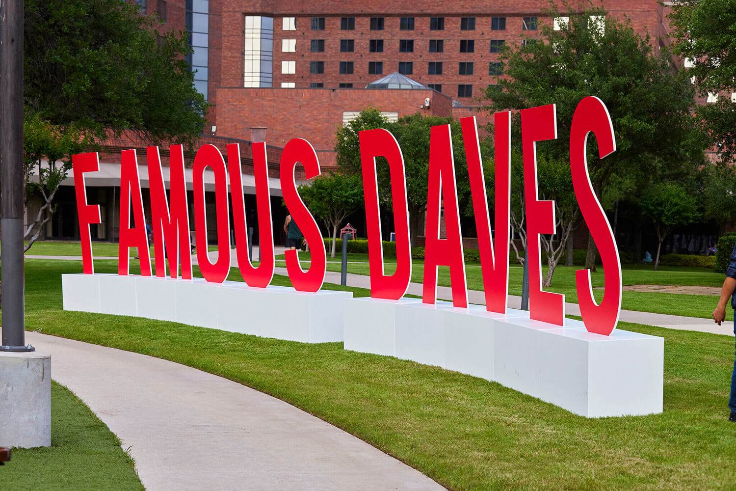 A bold red “Famous Dave’s” sign displayed on a white base outdoors at the 2024 BBQ Holdings Conference in Dallas, TX, attended by the Triple A Group. The backdrop features a brick building and a landscaped walkway.