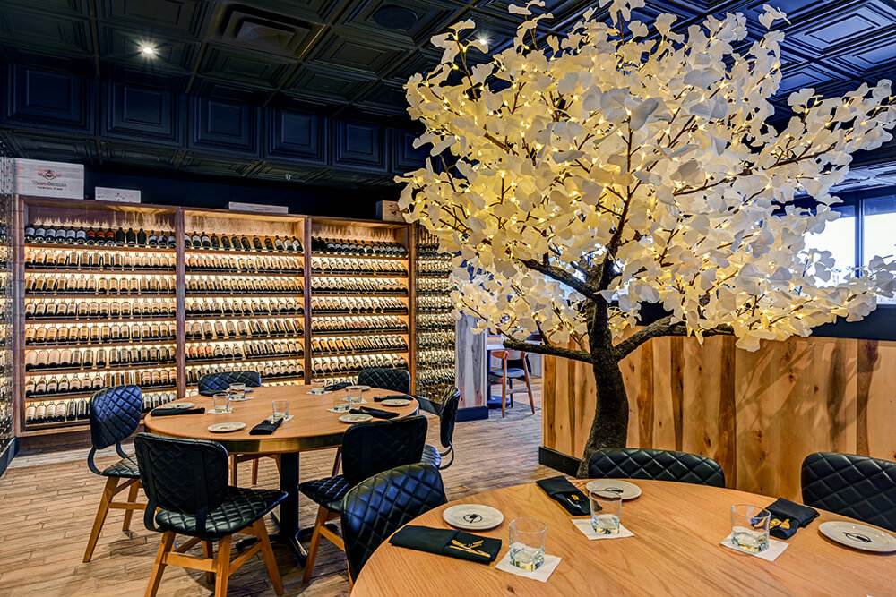 Elegant dining area featuring a backlit artificial tree with white leaves, surrounded by round wooden tables set for guests. A full wall of wine bottles in wooden racks creates a warm, inviting atmosphere ideal for upscale dining and special gatherings.