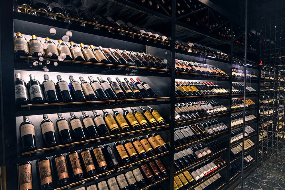 Extensive wine cellar featuring rows of premium wine bottles neatly arranged on modern black shelving, illuminated with soft lighting to highlight a diverse selection of labels and vintages.