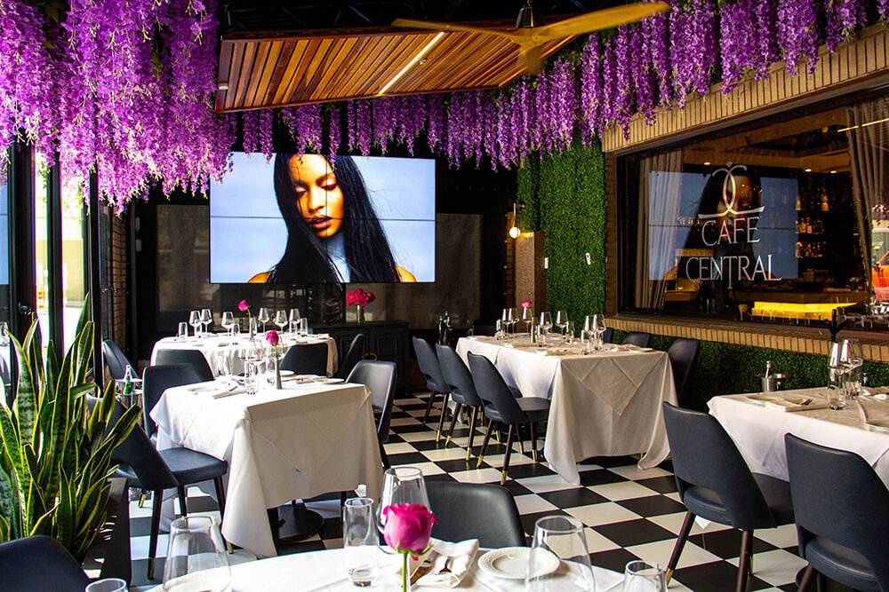 Interior view of Café Central’s elegant dining area featuring checkered floors, white tablecloths, and purple wisteria hanging from the ceiling. A digital art display and lush green wall create a stylish, modern ambiance.