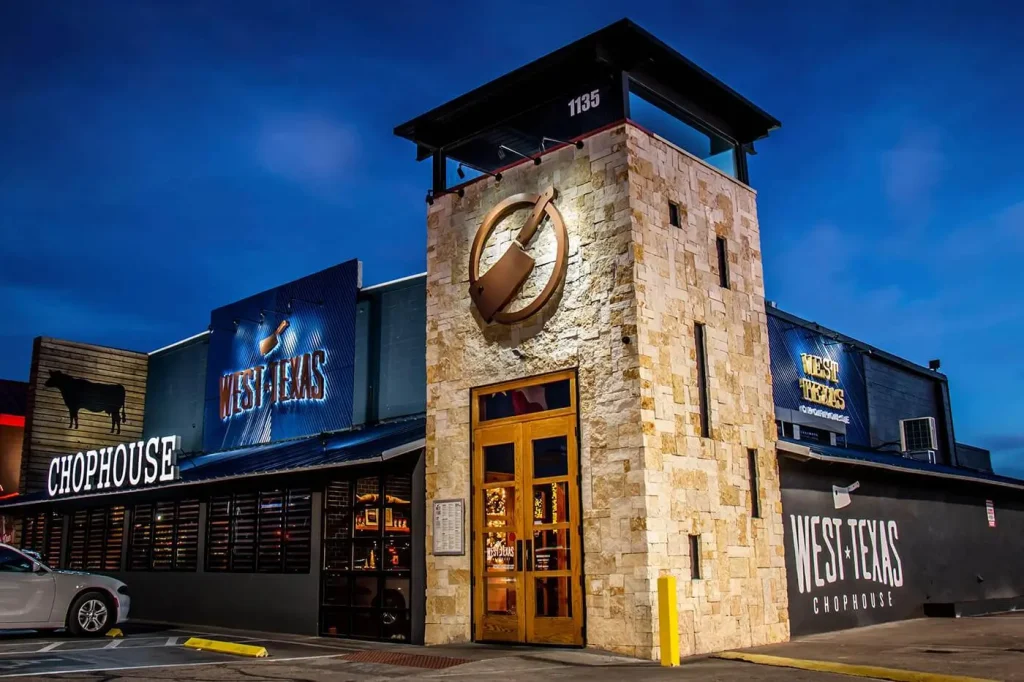 West Texas Chophouse restaurant exterior at night, featuring modern rustic architecture with stone tower, branded signage, and warm interior lighting — inviting guests to a premier Texas-style steakhouse dining experience.